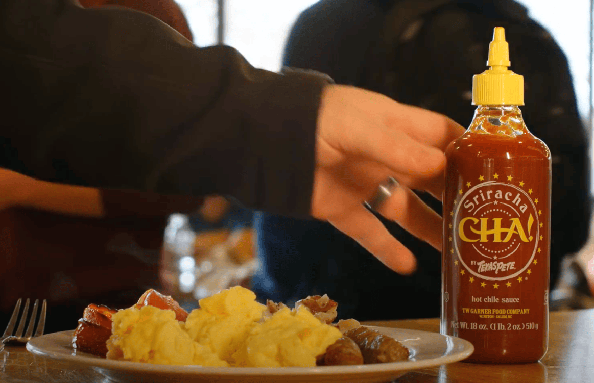 College breakfast bar and plate with a human hand reaching for a bottle of Cha Sriracha by Texas Pete